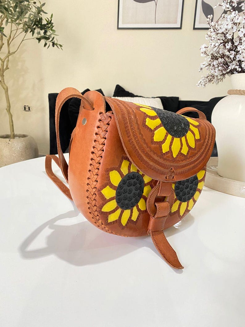 Handcrafted Mexican leather crossbody bag modeled by a woman in a rust sleeveless top and denim jeans. The bag is tan brown with hand-tooled sunflower details in bright yellow and black, featuring a curved flap, rustic stitching, and an adjustable leather strap. A unique bohemian accessory perfect for adding handmade floral charm and artisan craftsmanship to everyday outfits or travel looks.