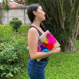 Handmade palm clutch featuring a rectangular woven palm leaf body, decorated with vibrant multicolor tassels and hand-painted pine cone flowers in pink, yellow, turquoise, purple, orange, and red. The center strip has a colorful embroidered textile accent, creating a unique artisan boho design. Perfect as a statement accessory for festivals, summer parties, vacations, or as a one-of-a-kind handmade gift for women.