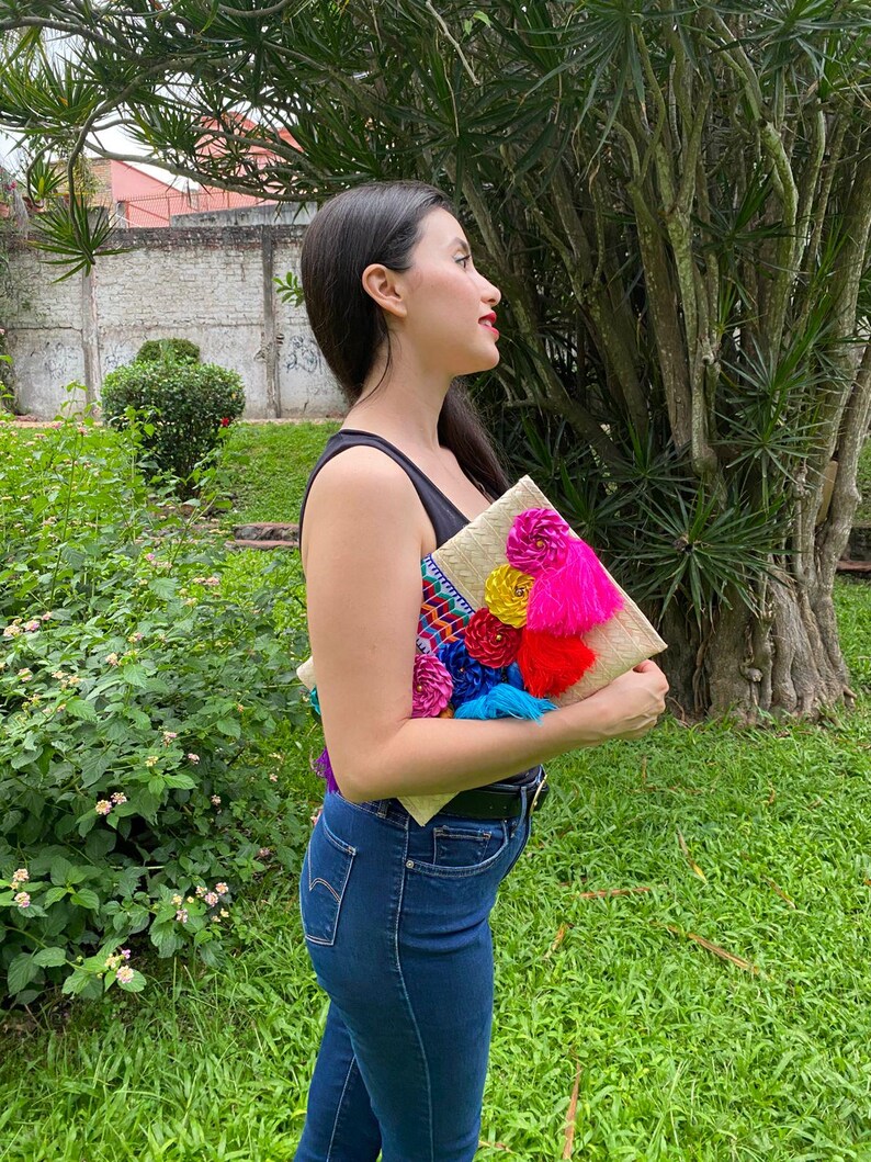 Handmade palm clutch featuring a rectangular woven palm leaf body, decorated with vibrant multicolor tassels and hand-painted pine cone flowers in pink, yellow, turquoise, purple, orange, and red. The center strip has a colorful embroidered textile accent, creating a unique artisan boho design. Perfect as a statement accessory for festivals, summer parties, vacations, or as a one-of-a-kind handmade gift for women.