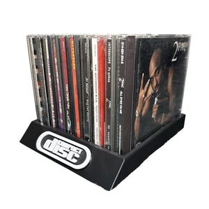 May include: A black plastic display rack holding a collection of music CDs, including albums by 2Pac, The Notorious B.I.G., and Pearl Jam. The front of the rack features a white and black Compact Disc logo. The CDs are arranged vertically in a row.