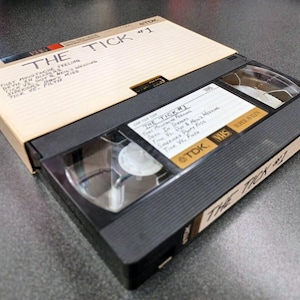 Può includere: Un nastro VHS nero con l'etichetta "The Tick #1" in bianco. Il nastro è etichettato con il titolo "The Tick #1" e il titolo dell'episodio "That Mustache Feeling". Il nastro è un nastro VHS TDK.