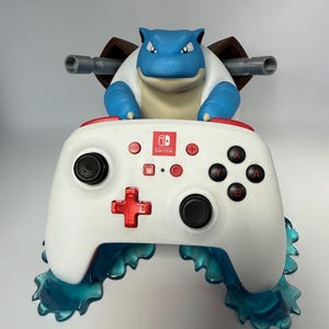 May include: A blue and white Blastoise character figure holding a white Nintendo Switch controller with red accents. The controller features black buttons, a red directional pad, and the Nintendo Switch logo. The base is made of translucent blue water.