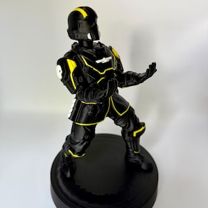 May include: A black and yellow figurine of a soldier in tactical armor standing on a circular base. The figure wears a helmet with a yellow stripe, a chest plate with a white emblem, and armor accents on the shoulders, arms, and legs.