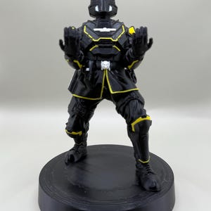 May include: A black figurine of a futuristic soldier with yellow accents. The figure is posed with arms raised, wearing a helmet and armor. It stands on a black circular base, showcasing intricate details and a dynamic design.