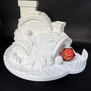 May include: A white 3D printed hobbit-style house sculpture with two chimneys, rounded doors, and a stone path. A translucent red twenty-sided die with gold-colored internal details sits in the open entrance of the lower dwelling section.