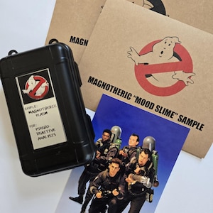 May include: A black case with a Ghostbusters logo and text, alongside brown paper cards with the Ghostbusters logo and text, and a blue card with a photo of the Ghostbusters team. The case reads "SAMPLE: MAGNOTHERIC PLASM".