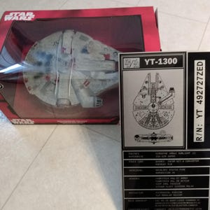 May include: A Star Wars Millennium Falcon model in a red box. The model is grey and white with red accents. A black and white information card with the text "YT-1300" and "R/N: YT 492727ZED" is next to the box.