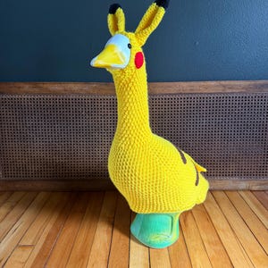 May include: A yellow crocheted goose head cover with black-tipped ears, a white and yellow beak, and red cheek accents. The goose head cover is on a green base. The item is designed to fit over a goose.