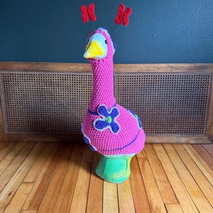 May include: A goose figurine wearing a bright pink crocheted cover with blue and green flower appliqués. The goose has a yellow beak and two red crocheted antennae attached to its head with thin wires, creating a colorful and textured appearance.