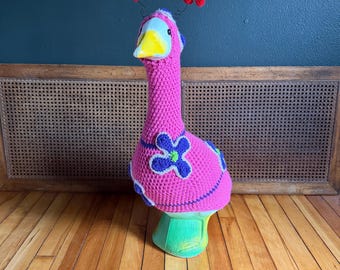 Könnte beinhalten: Eine Gänsefigur, die einen leuchtend pinkfarbenen, gehäkelten Überzug mit blauen und grünen Blumenapplikationen trägt. Die Gans hat einen gelben Schnabel und zwei rote, gehäkelte Antennen, die mit dünnen Drähten am Kopf befestigt sind.