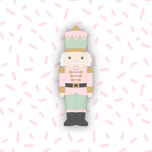May include: A pink and green nutcracker cookie cutter with a gold crown and accents. The nutcracker has a white moustache and hair, and black boots. The background is white with pink sprinkles.