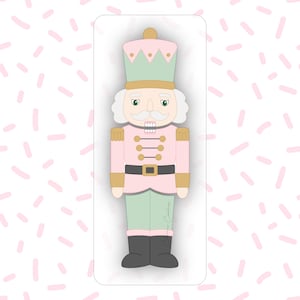 May include: A pink, green, and gold nutcracker cookie cutter. The cutter depicts a nutcracker soldier with a hat, uniform, and boots. The background is white with pink sprinkles.