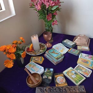 Puede incluir: Un mantel de mesa morado con una variedad de cartas de tarot, una vela blanca, un cáliz de cobre, un porta incienso de madera, un cuenco cantor y dos ramos de flores.
