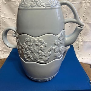 May include: A three-tiered ceramic teapot in a light blue color with an intricate floral design. The teapot has a handle and a spout.