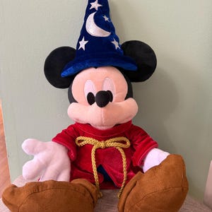 K&ouml;nnte beinhalten: Ein Mickey Mouse Pl&uuml;schtier als Zauberer verkleidet, mit einem blauen Hut mit Mond und Sternen. Er hat eine rote Robe mit goldener Kordel, schwarze Ohren und braune Schuhe. Das Spielzeug sitzt auf einem Stuhl.