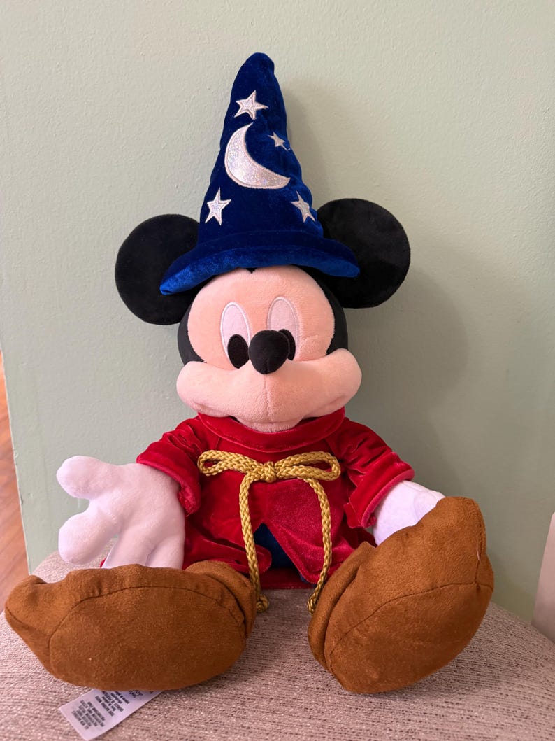 K&ouml;nnte beinhalten: Ein Mickey Mouse Pl&uuml;schtier als Zauberer verkleidet, mit einem blauen Hut mit Mond und Sternen. Er hat eine rote Robe mit goldener Kordel, schwarze Ohren und braune Schuhe. Das Spielzeug sitzt auf einem Stuhl.