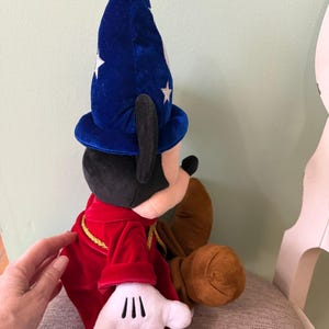 K&ouml;nnte beinhalten: Ein Mickey Mouse Pl&uuml;schtier, verkleidet als Zauberlehrling aus Fantasia. Mickey tr&auml;gt einen blauen Samt-Zauberhut mit wei&szlig;en Sternen, einen roten Samtumhang mit goldenen Verzierungen und wei&szlig;e Handschuhe. Das Spielzeug sitzt.