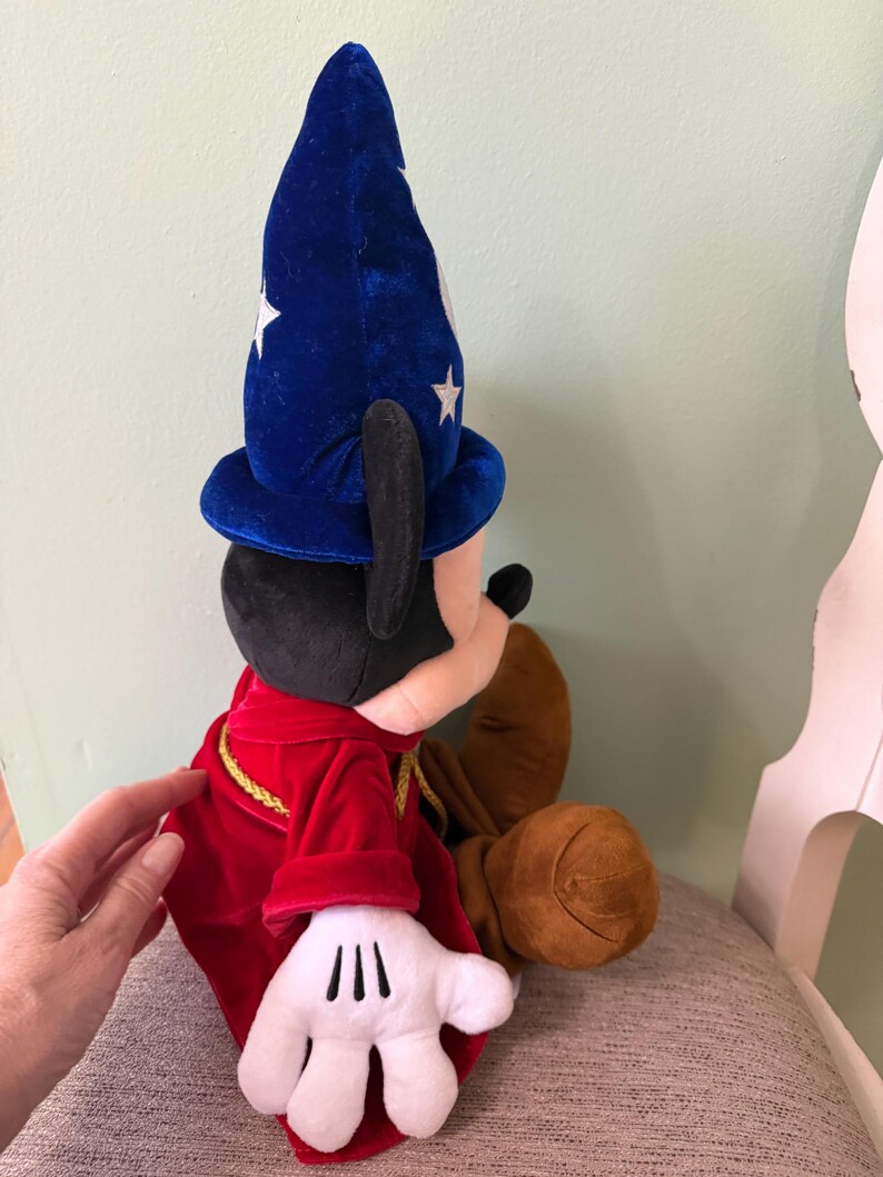 K&ouml;nnte beinhalten: Ein Mickey Mouse Pl&uuml;schtier, verkleidet als Zauberlehrling aus Fantasia. Mickey tr&auml;gt einen blauen Samt-Zauberhut mit wei&szlig;en Sternen, einen roten Samtumhang mit goldenen Verzierungen und wei&szlig;e Handschuhe. Das Spielzeug sitzt.