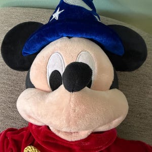 K&ouml;nnte beinhalten: Ein Mickey Mouse-Pl&uuml;schtier, das als Zauberer verkleidet ist. Mickey tr&auml;gt einen blauen Samt-Zauberhut mit silbernen Sternen und eine rote Samt-Robe mit einem goldenen Seilband. Das Spielzeug hat schwarze Ohren, ein rosa Gesicht und eine schwarze Nase.