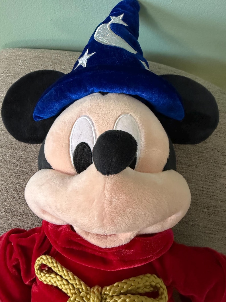 K&ouml;nnte beinhalten: Ein Mickey Mouse-Pl&uuml;schtier, das als Zauberer verkleidet ist. Mickey tr&auml;gt einen blauen Samt-Zauberhut mit silbernen Sternen und eine rote Samt-Robe mit einem goldenen Seilband. Das Spielzeug hat schwarze Ohren, ein rosa Gesicht und eine schwarze Nase.