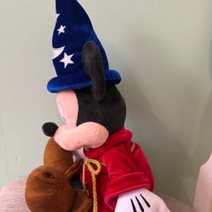 K&ouml;nnte beinhalten: Ein Pl&uuml;sch-Mickey Mouse als Zauberer, mit einem blauen Samthut mit wei&szlig;en Sternen und einem roten Samtgewand. Das Pl&uuml;schtier hat wei&szlig;e Handschuhe und braune Stiefel. Die goldene Kordel verleiht dem Outfit einen Hauch von Eleganz.