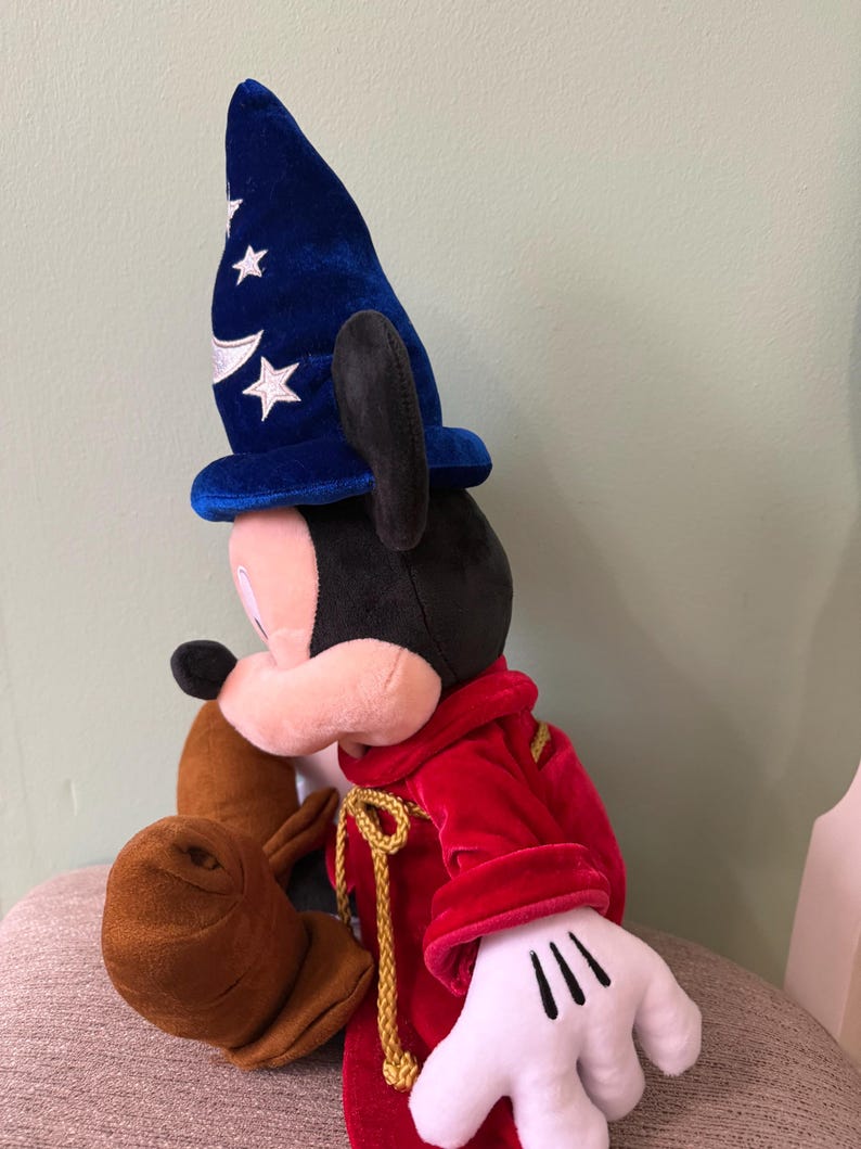K&ouml;nnte beinhalten: Ein Pl&uuml;sch-Mickey Mouse als Zauberer, mit einem blauen Samthut mit wei&szlig;en Sternen und einem roten Samtgewand. Das Pl&uuml;schtier hat wei&szlig;e Handschuhe und braune Stiefel. Die goldene Kordel verleiht dem Outfit einen Hauch von Eleganz.