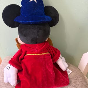 K&ouml;nnte beinhalten: Ein Mickey Mouse-Pl&uuml;schtier, das als Zauberer verkleidet ist und einen blauen Samthut mit einem wei&szlig;en Stern tr&auml;gt. Das Spielzeug hat eine rote Robe mit goldenem Besatz und wei&szlig;e Handschuhe. Das Spielzeug sitzt auf einer gemusterten Oberfl&auml;che.