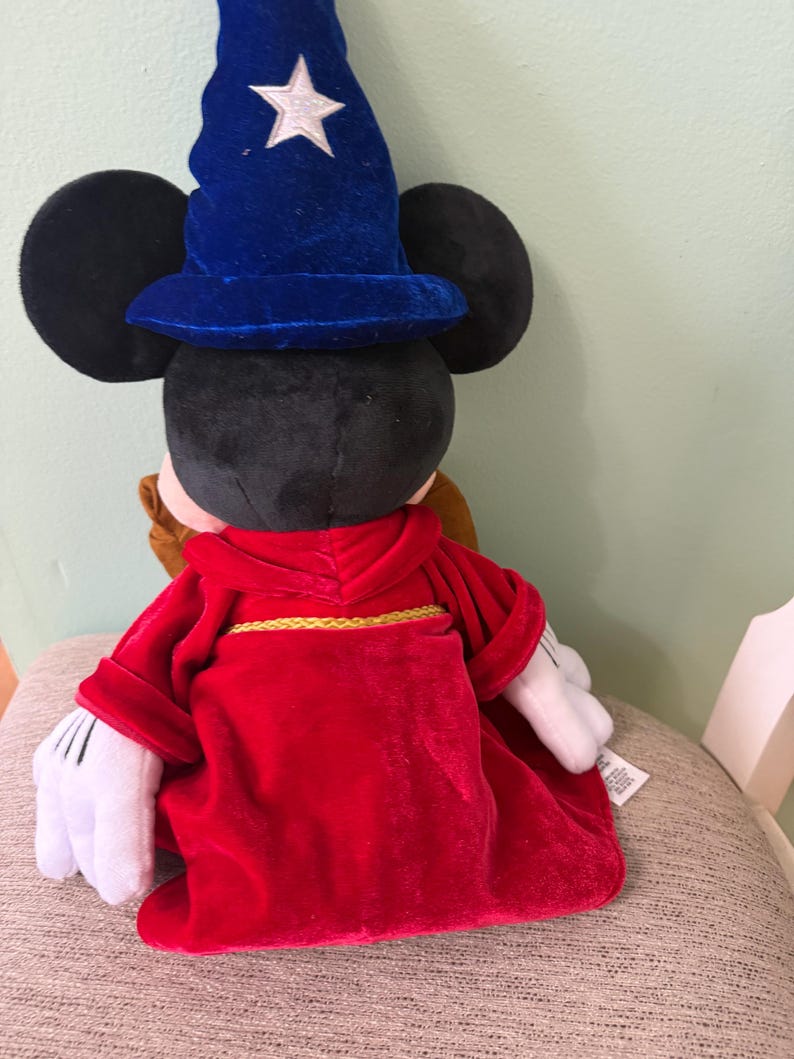 K&ouml;nnte beinhalten: Ein Mickey Mouse-Pl&uuml;schtier, das als Zauberer verkleidet ist und einen blauen Samthut mit einem wei&szlig;en Stern tr&auml;gt. Das Spielzeug hat eine rote Robe mit goldenem Besatz und wei&szlig;e Handschuhe. Das Spielzeug sitzt auf einer gemusterten Oberfl&auml;che.