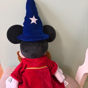 K&ouml;nnte beinhalten: Ein Mickey Mouse-Pl&uuml;schtier, das als Zauberer verkleidet ist und einen blauen Samthut mit einem wei&szlig;en Stern und eine rote Robe mit goldenem Besatz tr&auml;gt. Das Spielzeug hat schwarze Ohren und wei&szlig;e Handschuhe.