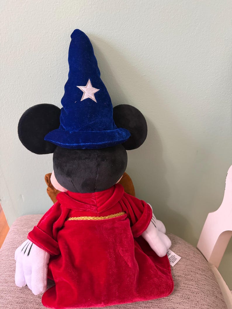 K&ouml;nnte beinhalten: Ein Mickey Mouse-Pl&uuml;schtier, das als Zauberer verkleidet ist und einen blauen Samthut mit einem wei&szlig;en Stern und eine rote Robe mit goldenem Besatz tr&auml;gt. Das Spielzeug hat schwarze Ohren und wei&szlig;e Handschuhe.