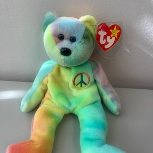 Puede incluir: Un oso de peluche tie-dye con un signo de paz en el pecho. El oso es verde, amarillo, naranja y azul. El oso tiene una etiqueta Ty unida a su oreja.