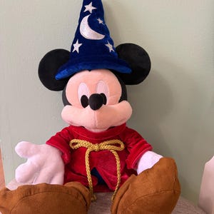 K&ouml;nnte beinhalten: Ein Mickey Mouse Pl&uuml;schtier als Zauberer verkleidet, mit einem blauen Hut mit wei&szlig;en Sternen und einem Halbmond. Er hat eine rote Robe mit einem goldenen Seilbinder und braunen Schuhen. Das Pl&uuml;schtier sitzt auf einem Stuhl.