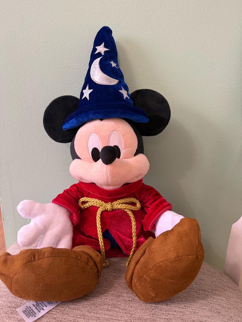 K&ouml;nnte beinhalten: Ein Mickey Mouse Pl&uuml;schtier als Zauberer verkleidet, mit einem blauen Hut mit wei&szlig;en Sternen und einem Halbmond. Er hat eine rote Robe mit einem goldenen Seilbinder und braunen Schuhen. Das Pl&uuml;schtier sitzt auf einem Stuhl.