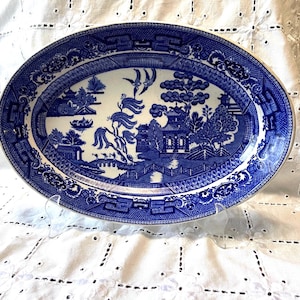 May include: Oval blue and white ceramic platter with a Willow pattern design. The platter features a detailed landscape scene with pagodas, bridges, and trees. The rim has a decorative border. This vintage serving dish is ideal for displaying or serving food.