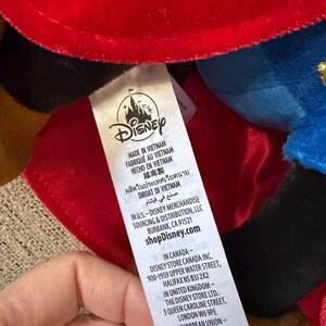 K&ouml;nnte beinhalten: Ein Disney-Produktetikett mit dem Disney-Logo und Text in mehreren Sprachen, darunter Deutsch. Das Etikett gibt an, dass das Produkt in Vietnam hergestellt wurde und die Website shopDisney.com.