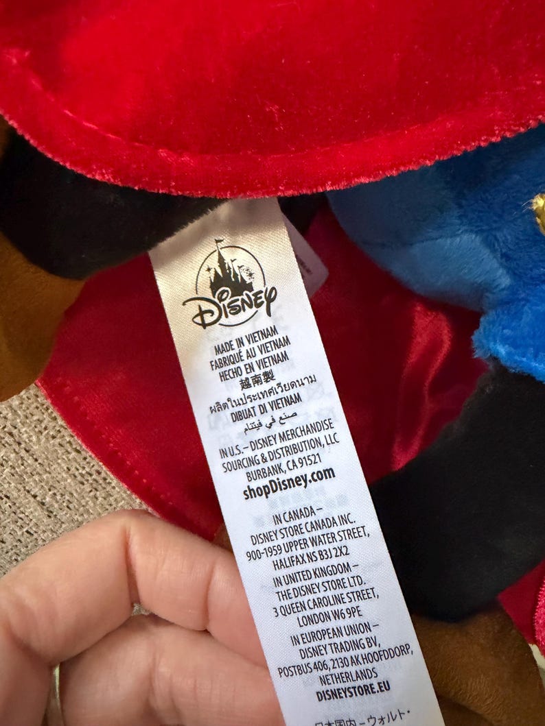 K&ouml;nnte beinhalten: Ein Disney-Produktetikett mit dem Disney-Logo und Text in mehreren Sprachen, darunter Deutsch. Das Etikett gibt an, dass das Produkt in Vietnam hergestellt wurde und die Website shopDisney.com.