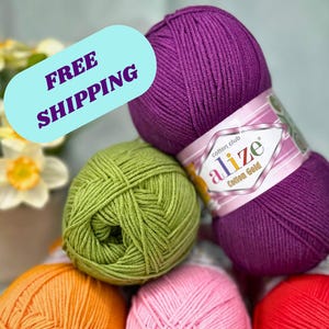 以下が含まれることがあります： 紫、緑、オレンジ、ピンク、赤の色合いのカラフルな毛糸玉のコレクション。毛糸は山のように配置され、紫色の毛糸玉にはラベルが見えます。画像は柔らかく、拡散した照明です。