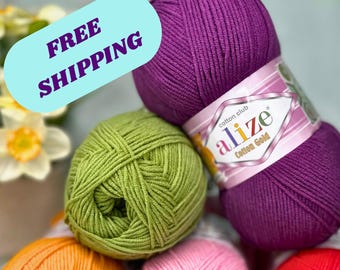 May include: A collection of colorful yarn balls in shades of purple, green, orange, pink, and red. The yarn is arranged in a pile, with a label visible on the purple skein. The image has a soft, diffused lighting.