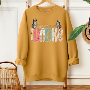 Puede incluir: Sudadera de color amarillo mostaza con las palabras "give THANKS" en letras coloridas y estampadas. Dos mariposas están encima de la palabra "give". Las letras están decoradas con estampados florales, de rayas y de lunares.