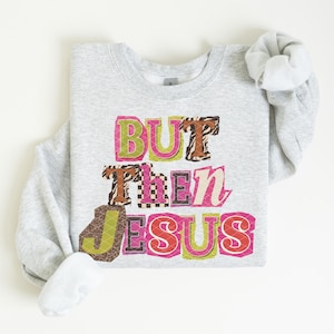 May include: A light gray sweatshirt with the text "BUT THEN JESUS" in a colorful, patchwork-style design. The letters feature various patterns, including animal print and checkered designs, with pink, green, and orange accents.
