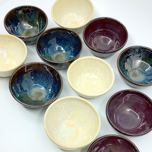 May include: A collection of ten small ceramic bowls in various colors, including cream, blue, brown, and purple. The bowls are all round and have a smooth, glossy finish.