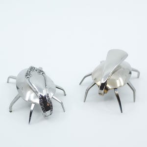 May include: Two silver metal bugs made from spoons. The bugs have six legs and are posed on a white surface.