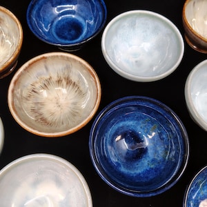 May include: An overhead shot showcases a collection of ceramic bowls in various shades of blue, white, and brown. The bowls have a glossy finish and unique patterns, with some featuring speckled or marbled designs. The bowls are arranged on a black surface.