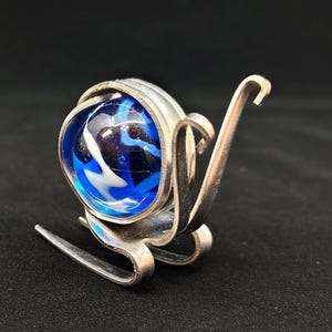 May include: A unique, handcrafted silver-toned metal sculpture featuring a blue and white swirled glass marble. The sculpture has a curved, abstract design with a polished finish, set against a dark background.