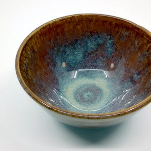 May include: A handmade ceramic bowl with a glossy finish. The interior features a blend of teal, brown, and cream colors, creating a unique, swirling pattern. The rim is a darker brown, and the bowl has a smooth, rounded shape.