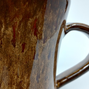 May include: A brown ceramic mug with a mottled glaze and a large handle. The glaze has a reddish-brown color with streaks of a lighter brown.