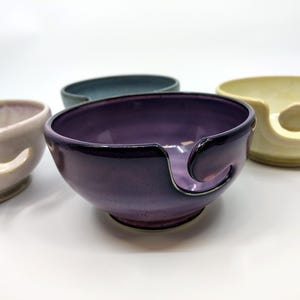 May include: A collection of ceramic yarn bowls in various colors, including purple, blue, and cream. Each bowl has a curved slot for yarn and a rounded shape. The bowls are arranged on a white surface.