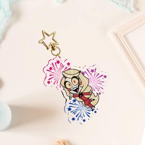 May include: An acrylic keychain featuring a cartoon character with blonde hair, red cheeks, and a red suit, surrounded by pink and blue firework illustrations. The keychain is attached to a gold-colored star-shaped metal clasp at the top.