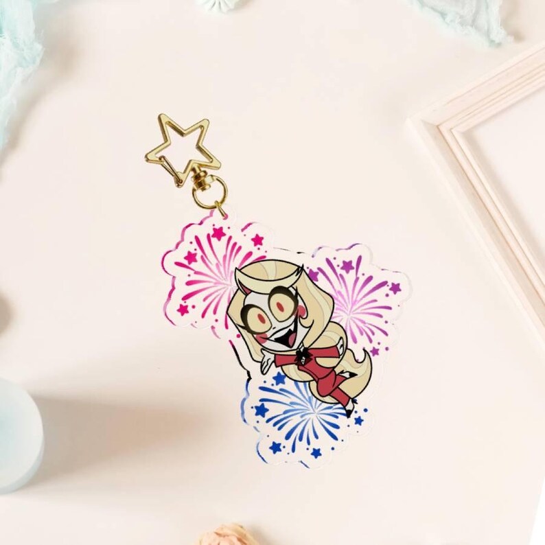 May include: An acrylic keychain featuring a cartoon character with blonde hair, red cheeks, and a red suit, surrounded by pink and blue firework illustrations. The keychain is attached to a gold-colored star-shaped metal clasp at the top.