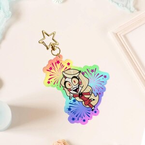 May include: An acrylic keyring featuring a cartoon character with blonde hair and a red outfit, set against a vibrant rainbow-coloured background with firework and star patterns. The charm is attached to a gold-coloured star-shaped metal clasp.